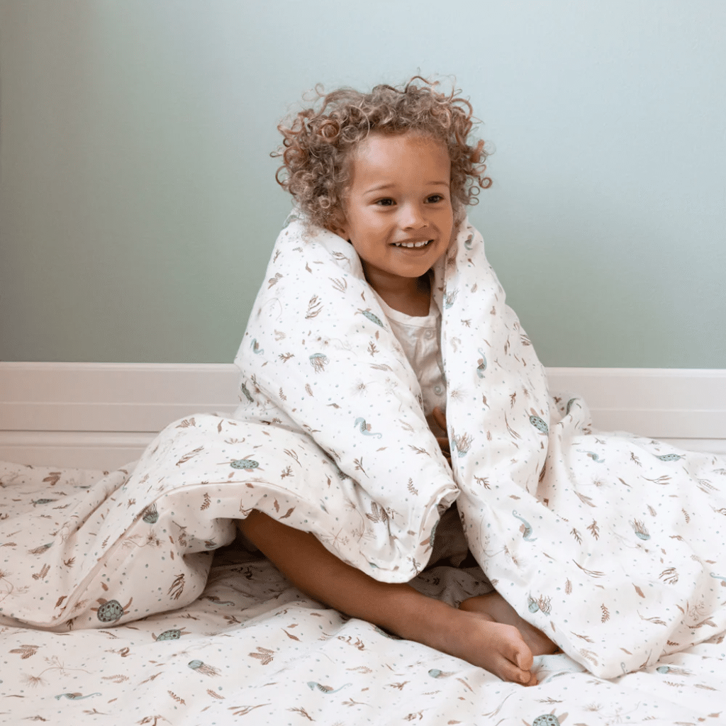 a child sitting on a bed covered with a blanket Cam cam Sengetøj, Junior, 100x140cm - GOTS sea garden