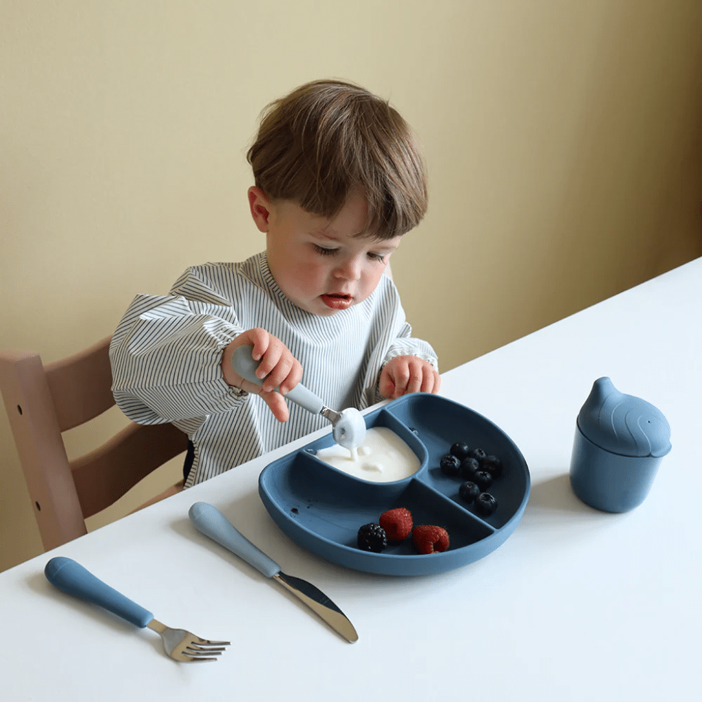 a child eating with a spoon and a plate of food. cam cam Hagesmæk med Ærmer - Classic Stripes Blue